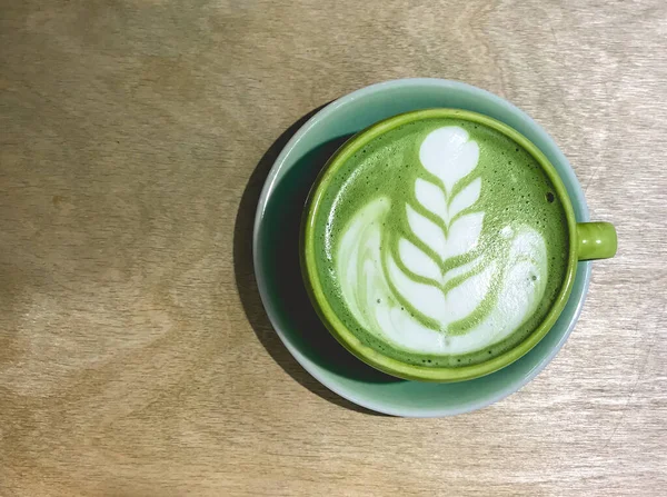 Green cup with traditional japanese matcha latte green tea with a leaf and flower design in the foam over a light blue saucer in a wood table seen from above