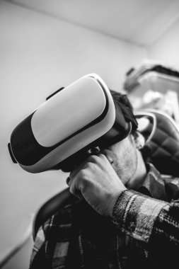 Ekose gömlekli yakışıklı ve ilgili bir genç adam, evdeki oyun sandalyesinde sanal gerçeklik kaskıyla (VR gözlükleri) oynuyor.)