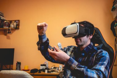 Yakışıklı ve ekose gömlekli genç Latin bir adam evde turuncu yatak odasındaki bilgisayar ekranının önünde sanal gerçeklik kaskıyla (VR gözlükle) oynuyor.