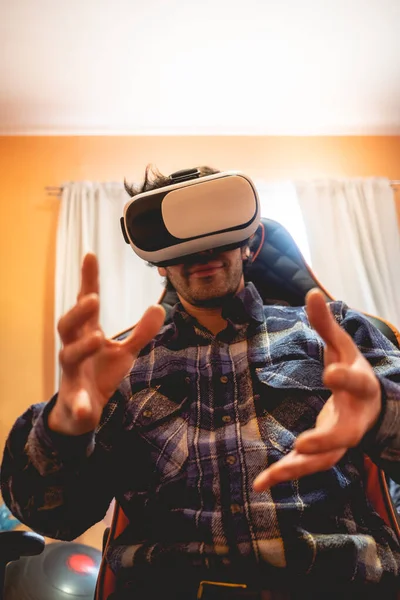 Handsome and happy young latino man with plaid shirt playing with virtual reality helmet (VR glasses) on a gamer chair with luminous window behind in orange bedroom on home