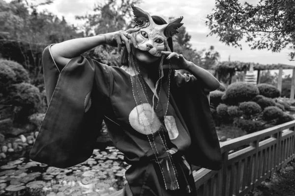 Portrait of sexy and young japanese girl with beautiful old traditional red kimono and handmade cat mask dancing in front of a pond in a japanese garden (in black and white)