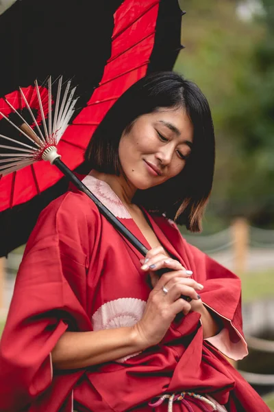 Portrait of sexy and young japanese girl with beautiful old traditional red kimono and handmade red and black paper umbrella in a japanese garden