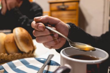 Closeup of hand of latino worker man holding a tea spoon with tea  and table with cup, bread and another man in the background