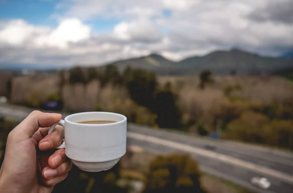 Hand holding a hot latte coffee in a white cup over a blurry background of a landscape with forest, mountains, blue sky and a highway