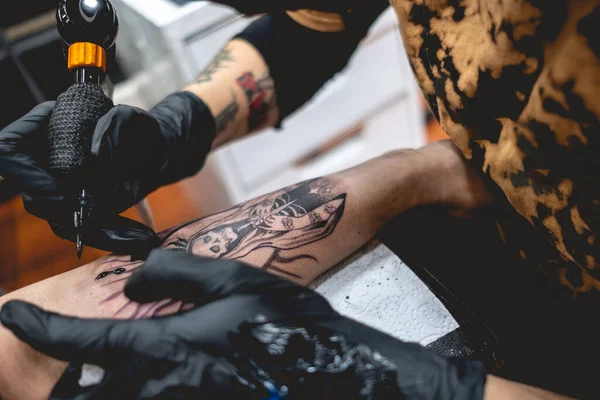 Detail of young tattoo artist girl with glasses and mask making a tattoo of \'La Santa Muerte\' (Our Lady of Holy Death) in the arm of a woman with tattoo machine