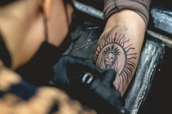 Detail of young tattoo artist girl with glasses and mask making a tattoo of \'La Santa Muerte\' (Our Lady of Holy Death) in the arm of a woman with tattoo machine