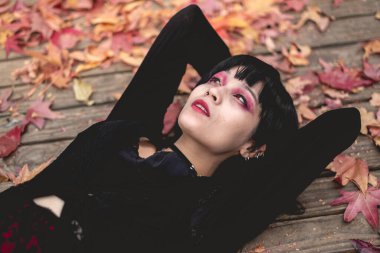 Siyah elbiseli, haçlı, seksi yüzlü ve kırmızı makyajlı genç ve sıska İspanyol gotik kız sonbahar ormanında kırmızı, turuncu ve sarı kuru yapraklarla bir ahşap platformda yatıyor.