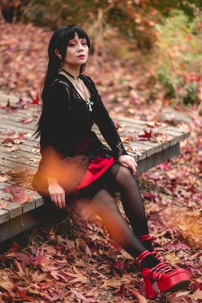 Young and skinny hispanic goth girl with black dress and red platforms shoes seated in a wood platform in the autumn forest with red, orange and yellow dry leaves