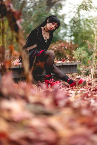 秋天的森林里 穿着黑色连衣裙和红色月台鞋的年轻瘦小的惊慌失措的女孩坐在一个木制平台上 有着红色 橙色和黄色的干叶 — 图库照片