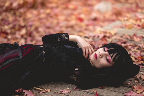 Young and skinny hispanic goth girl with black dress, cross, sexy face and red makeup lying in a wood platform in the autumn forest with red, orange and yellow dry leaves