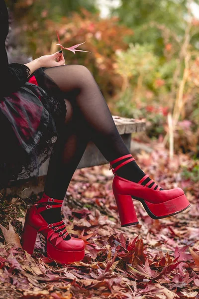 Young and skinny goth girl with black and red dress and red platforms shoes seated in a wood platform in the autumn forest holding red, orange and yellow dry leaves and no face