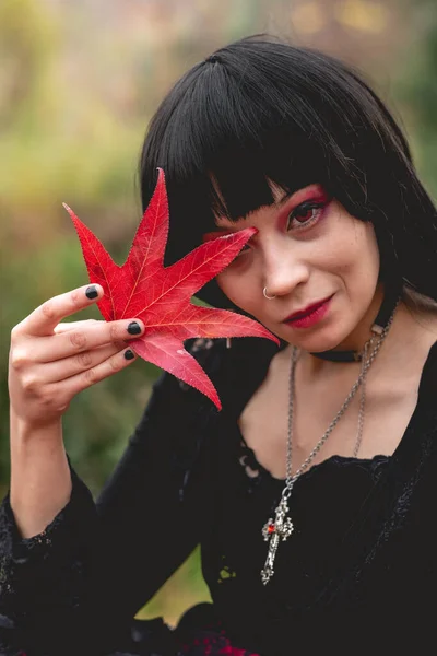 Young and skinny hispanic goth girl in the forest with black dress, cross, sexy face and red makeup holding a red and big dry leaf