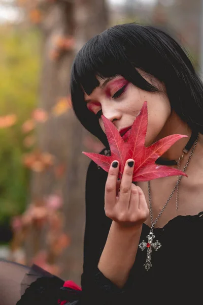 Young and skinny hispanic goth girl in the forest with black dress, cross, sexy face and red makeup holding a red and big dry leaf