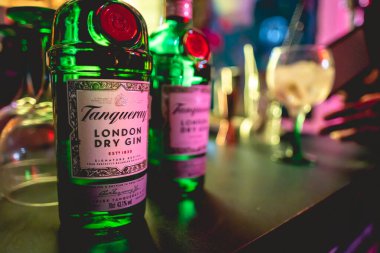 Tanqueray şişelerinin detayları. Kuru cin ve el. Gecenin bir yarısı renkli ışıklarla dolu bir barda arka planda kokteyl içiyorlar.