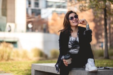 Genç, başarılı ve mutlu, güneş gözlüklü, defterli ve beyaz spor ayakkabılı Latin bir iş kadını gökdelenler arasındaki bir parkta kahve içiyor. 