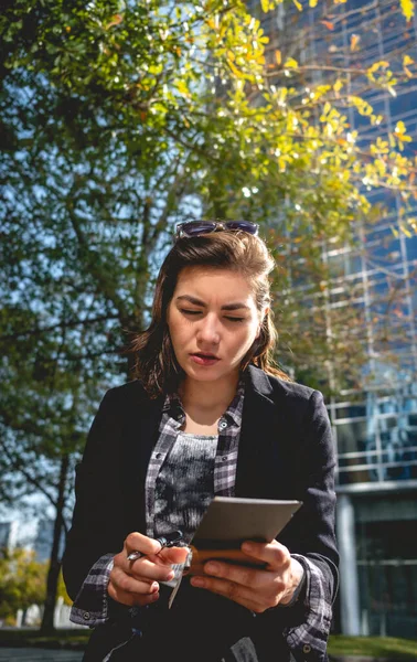 Young, successful and concentrated latina businesswoman with sunglasses and a cup of coffee taking notes of their ideas in a notebook in a park between skyscrapers