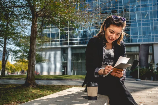 Young, successful and happy latina businesswoman with sunglasses and a cup of coffee taking notes of their ideas in a notebook in a park between skyscrapers