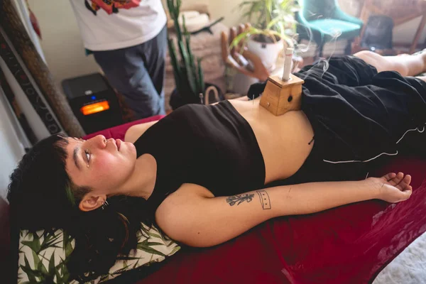 Herbal moxa stick in a smoke box over a teenager girl patient (moxibustion) with acupuncture needles in the skin and traditional chinese medicine doctor behind