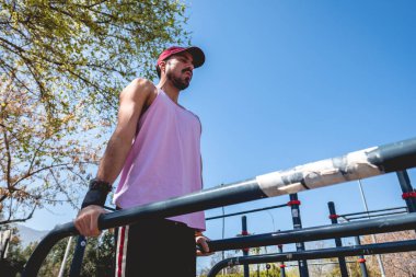 Yoğunlaşmış, sakallı, sağlıklı ve formda, mor tişörtlü ve kepli genç Latin bir adam güneşli bir günde bir sokak çalışma parkında jimnastik egzersizi yapıyor.