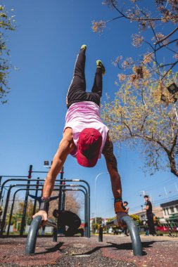 Sağlıklı ve formda kolunda ve omzunda dövmesi olan genç bir Latin adam, güneşli bir günde bir sokak çalışma parkında jimnastik (ters poz) yapıyor.