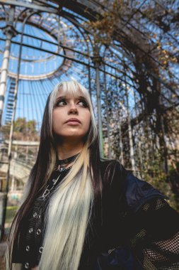 Terk edilmiş bir seranın içinde uzun sarı ve siyah saçlı, siyah elbiseli ve makyajlı genç ve zayıf bir manken.