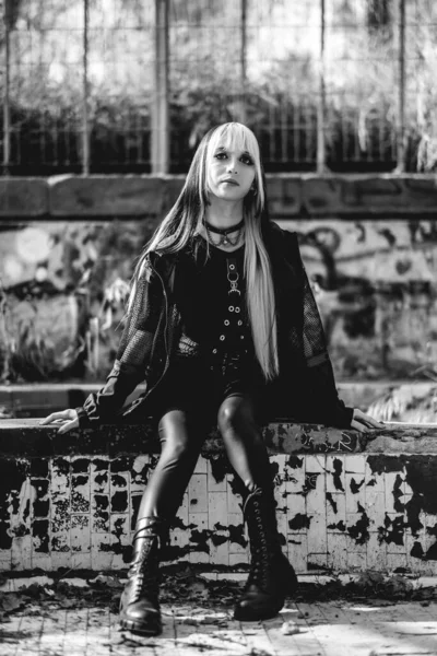 Young and skinny model with long blonde and black hair and black clothes and makeup sitting inside an old abandoned greenhouse (in black and white)