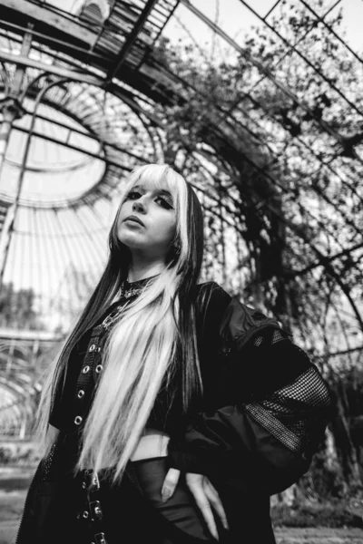 Young and skinny model with long blonde and black hair and black clothes and makeup posing inside an old abandoned greenhouse (in black and white)