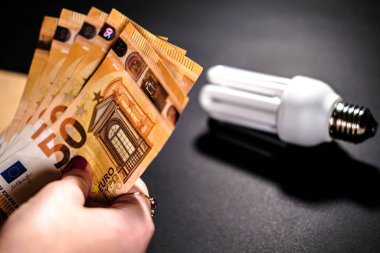 Arka planda bir avuç 50 avroluk banknot ve ampul tutan bir el resmi. Kavram: elektrik faturası zammı