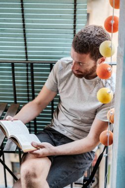 Vertical picture of red head beard man read an opened book at home. He enjoys the leisure time and good summer weather by the balcony. Colors light bulbs hanging and wooden blinders in the background