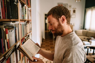 A red head male reading a book at home. He analyze and study literature for teaching and researching. He browse books in his personal library