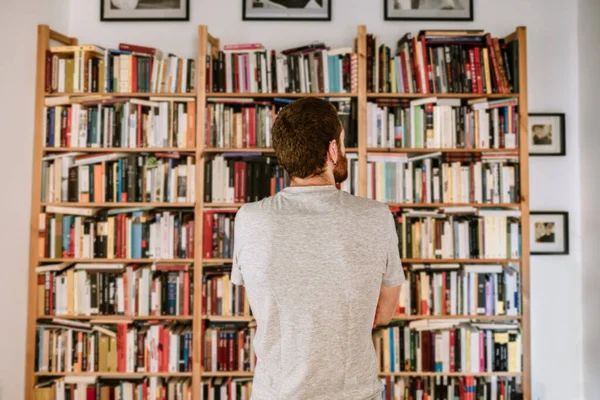 A red head observing his home library in his apartment. He is with his back turned. Red head bearded caucasian male. Thousands of books together. Books collection