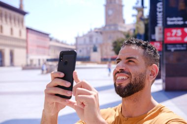 Happy and smiling arabic male taking a picture with his mobile phone. He is sightseeing Zaragoza in Spain and is at Plaza del Pilar. There is a christian cathedral in the background. Concept: tourism
