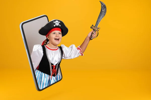 A little girl dressed as a pirate for Halloween holds a saber and looks from a smartphone. Child online on a mobile phone screen on a yellow background.