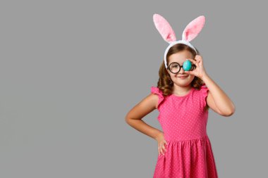 Paskalya yumurtasıyla gri arka planda küçük bir kızın portresi. Tavşan kulaklı ve gözlüklü neşeli çocuk.