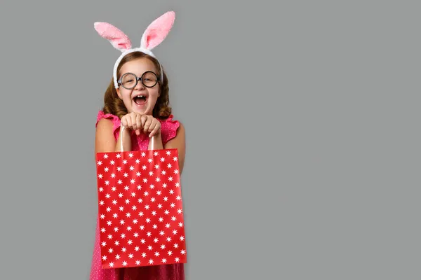 Mutlu Paskalyalar. Puantiyeli pembe elbiseli neşeli küçük bir kız elinde bir alışveriş çantası tutuyor. Gri arka planda şirin bir bebek..