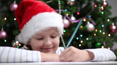 Sevimli çocuk, Noel Baba 'ya arka planda süslenmiş bir Noel ağacının ışıklarıyla bir mektup yazıyor..