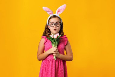 Kulaklı küçük bir kız ve sarı arka planda Paskalya tavşanı gözlüğü. Çocuk bir buket lale tutuyor..