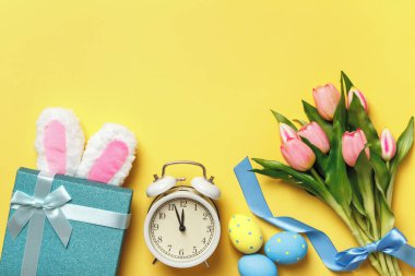 Paskalya yumurtaları çalar saat buketi, sarı arka planda tavşan kulaklı pembe hediye laleleri. Düz yatay kopyalama alanı üst görünümü.