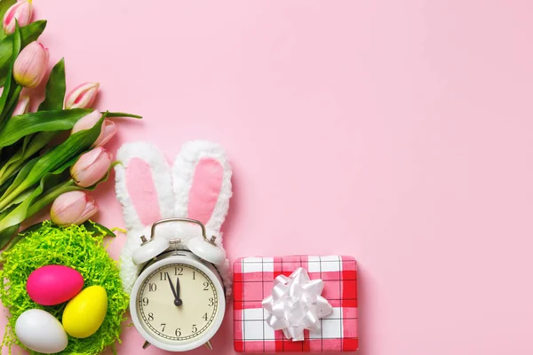 イースターカード 目覚まし時計ウサギの耳はピンクの背景に卵の花チューリップを贈っています フラットレイアウトコピースペーストップビュー — ストック写真