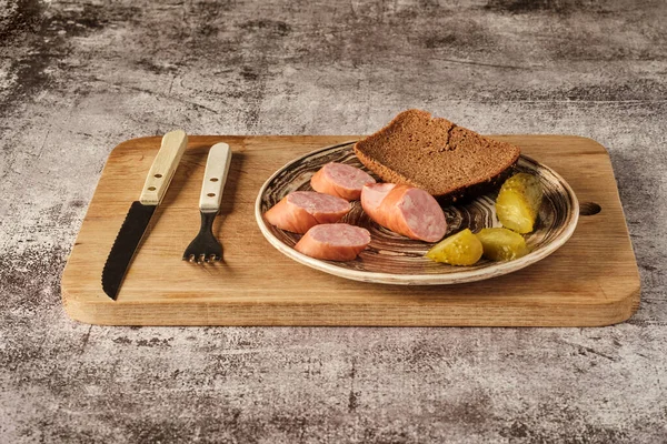 on a clay plate are pieces of sausage, pickles and bread, fork and knife, rustic style