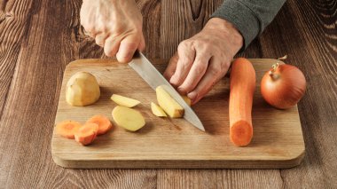 Bir adam mutfak tahtasına bıçakla patates kesiyor, havuç ve soğanın yanında.