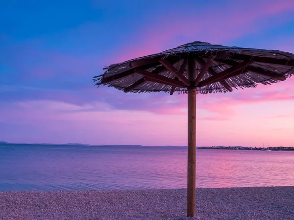 Beach umbrella, view of the beautiful blue and purple sunset, sky and straw beach umbrella. Perfect holiday concept. Travel and vacation. Beach on Vir Island, Croatia, Europa. Copy space.