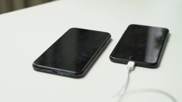 关闭两个黑色智能手机的视频 其中一个插在白桌上的白耳机上 — 图库视频影像