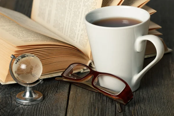 Warm tea cup on wooden table with old book, glasses and glass globe. — Stock Photo, Image