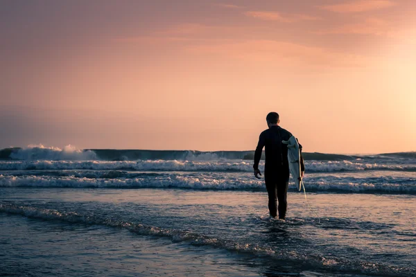 Mann surft im schwarzen Taucheranzug mit weißem Surfbrett am Strand bei Sonnenuntergang. — Stockfoto