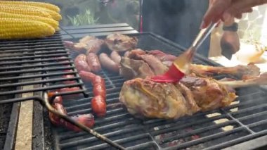 Şef kuzu budu ve domuz pirzolasına lezzetli sos ve sos koydu. Koçanında mısırla birlikte metal tepside ızgara yaptı. Sucuklu sucuk ve ızgarada lezzetli mısırlar pişiriyorum..