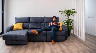 Neşeli yakışıklı adam evdeki kanepede uzanırken akıllı telefonunu kullanıyor. Kendine güveni tam sakallı adam akıllı telefonuyla evin kanepesinde daktilo çekiyor..