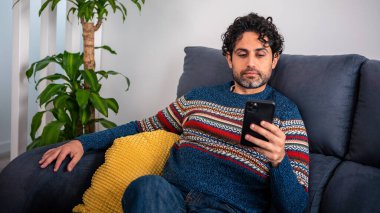 Evdeki kanepede otururken akıllı telefonunu kullanan neşeli yakışıklı bir adam. Kendine güveni tam sakallı adam akıllı telefonuyla evin kanepesinde daktilo çekiyor..