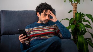 Akıllı telefon kullanıp güzel bir evde kanepede otururken nahoş bir acı hisseden yaşlı bir adamın portresi. Mutsuz Kafkasyalı adam telefon cihazıyla evin kanepesinde dinleniyor..