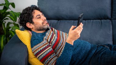 Akıllı telefonunu kullanarak gülümseyen ve evdeki kanepede uzanan mutlu yakışıklı adam. Kendine güveni tam sakallı bir adam. Evdeki koltukta rahatlıyor ve daktilo kullanıyor..